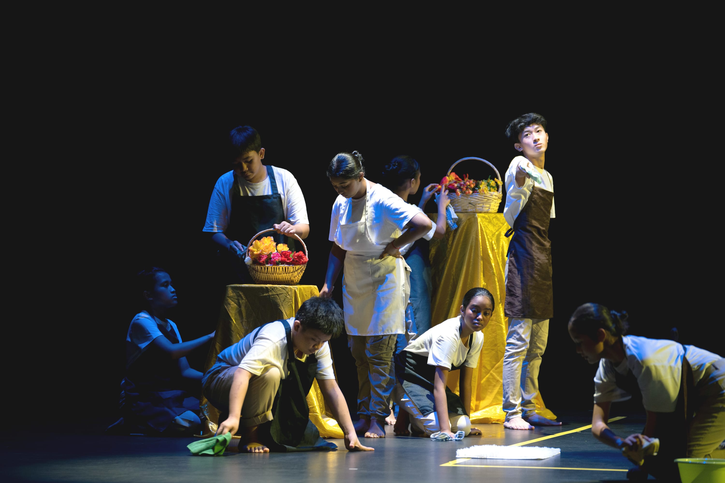 Young performers on a dark stage, some crouching and wiping the floor, others standing near tables with flower baskets.